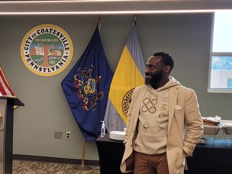 Ajené at a community gathering in Coatesville City Hall, beside the Pennsylvania and Coatesville flags and the city seal.