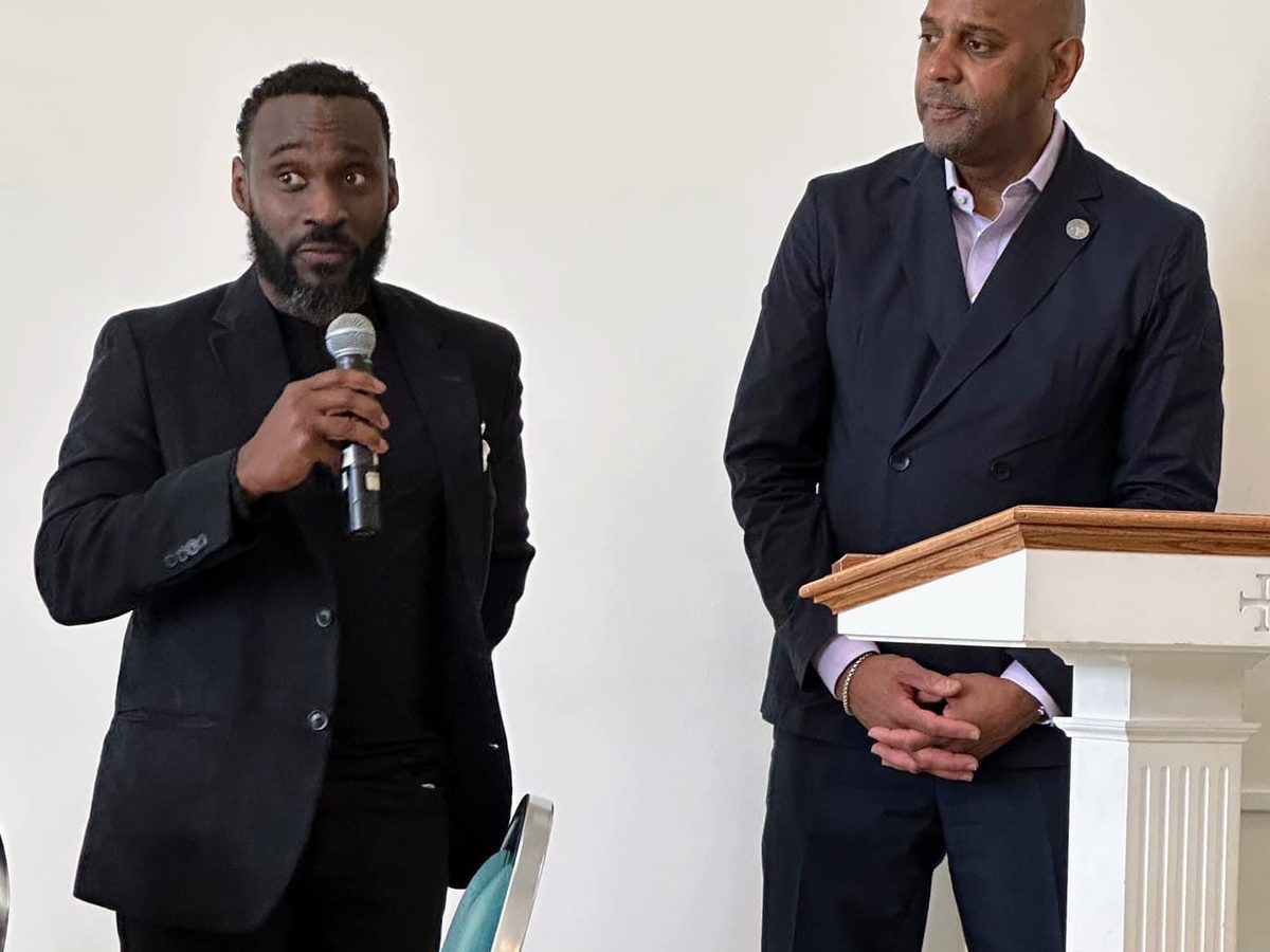 Ajené speaking with a microphone at a community meeting, standing beside a colleague at the podium.