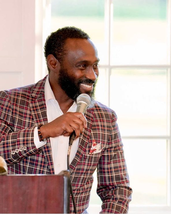 Ajené smiling with a microphone at a lectern, in a plaid blazer.