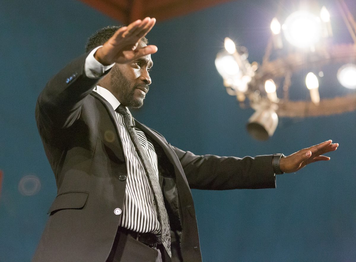 Ajené on stage as the Preacher in The Gospel at Colonus, arms outstretched beneath a candle chandelier.