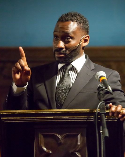 Ajené at the lectern in The Gospel at Colonus, one finger raised as he preaches.