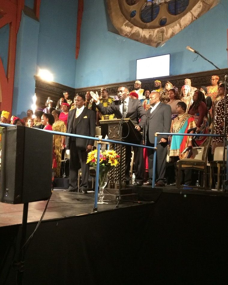 The full company of Coatesville Cultural Society's The Gospel at Colonus on stage at First Unitarian Church, Philadelphia, with Ajené at the lectern.
