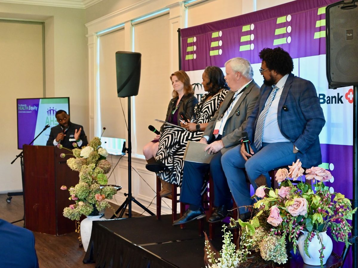 Ajené moderating a panel from the lectern at an Alliance for Health Equity event, with four panelists seated on stage.