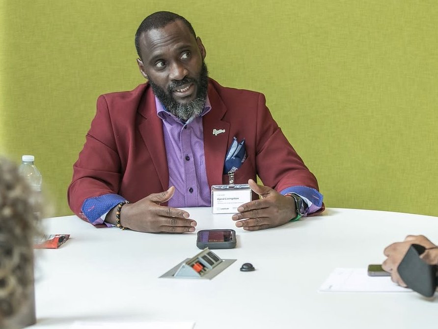 Ajené, in a red blazer and LeadLab name badge, talking with participants around a table during a LeadLab small-group session.