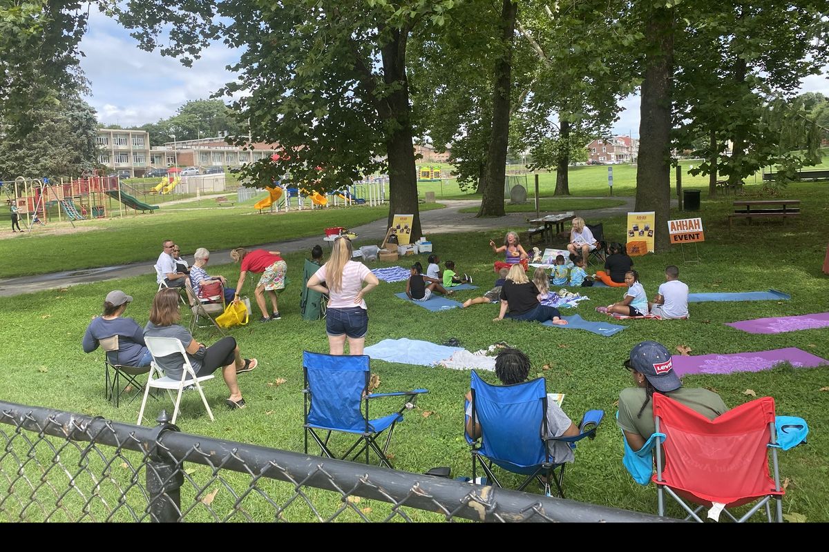 Children gathered on mats for storytelling under shade trees while families watch from lawn chairs at Sundays in the Park.