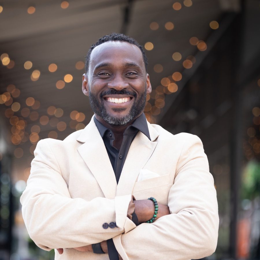 Ajené Livingston smiling, arms crossed, in a cream blazer under string lights.
