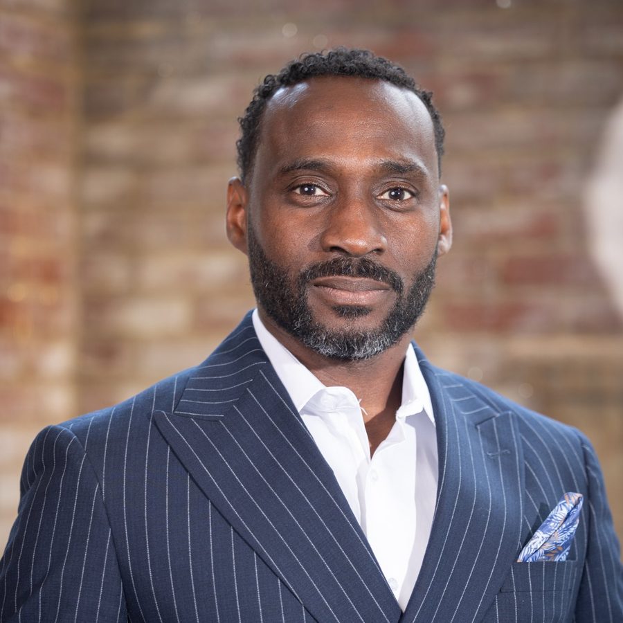 Portrait of Ajené Livingston in a navy pinstripe double-breasted suit against a brick wall.