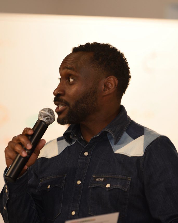 Ajené speaking into a microphone at a community event, holding a program card.